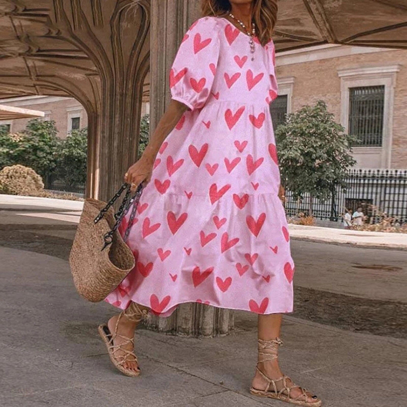 A woman wearing a pink midi dress with a red heart print and puff sleeves, carrying a straw bag.