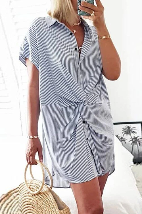 A woman in a blue and white striped shirt dress with a twist knot, holding a straw bag and a phone.