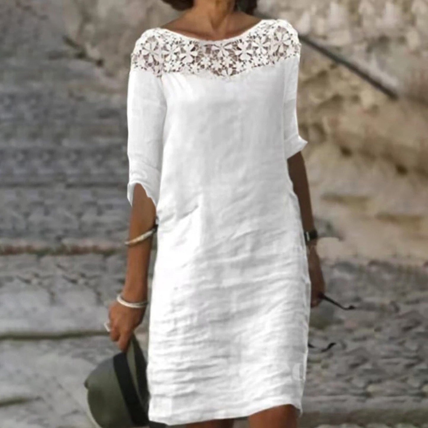 A woman wearing a white linen-like shift dress with a lace neckline and three-quarter sleeves, holding a hat and sunglasses.