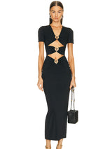 A woman stands facing forward wearing a black, floor-length dress with short sleeves and cutouts at the waist and chest, accented with gold rings. She holds a small black handbag and wears gold earrings.