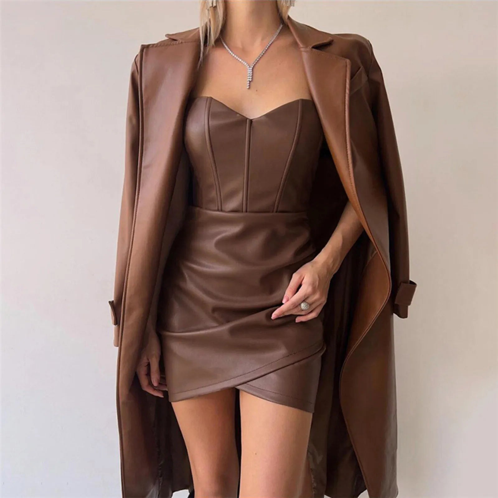 A woman wearing a brown strapless leather corset dress with a ruched skirt, layered under a brown leather trench coat.