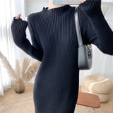 Close-up front view of a woman wearing a long-sleeved, black ribbed knit dress with a mock neck, holding a black handbag.