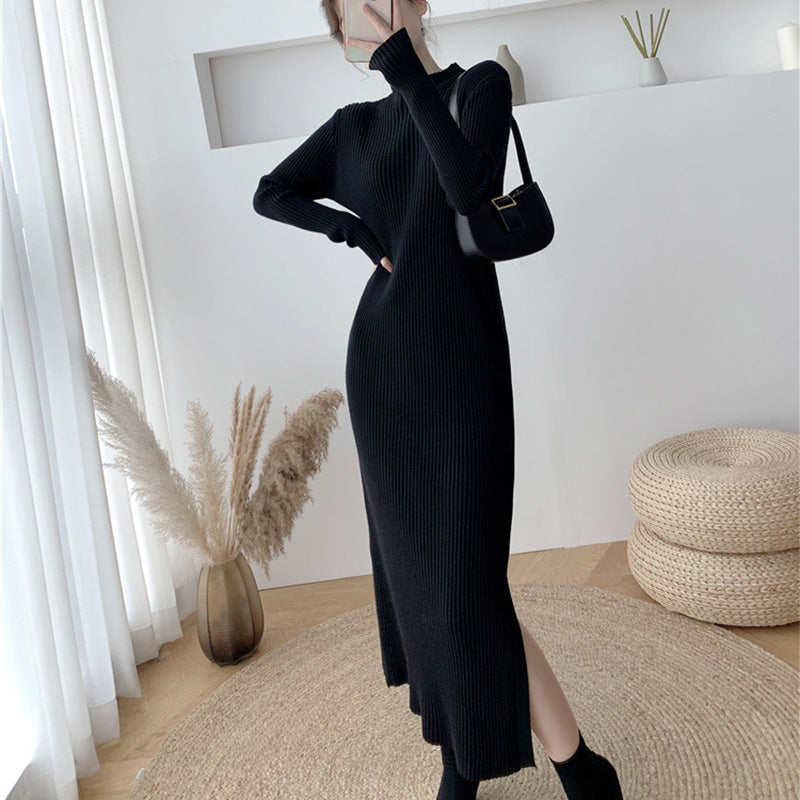 Woman wearing a long-sleeved, black ribbed knit dress with a side slit, black socks, and black pointed-toe shoes, holding a black handbag.