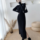 Woman wearing a long-sleeved, black ribbed knit dress with a side slit, black socks, and black pointed-toe shoes, holding a black handbag.