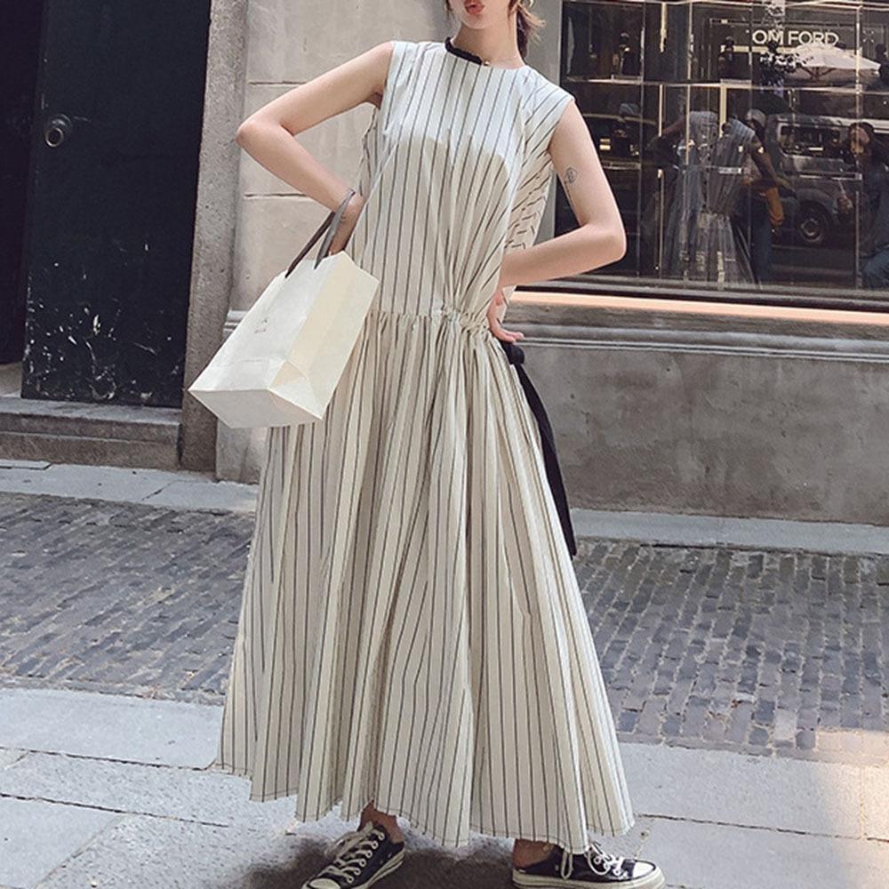 Woman wearing a long white sleeveless dress with black vertical stripes and a black ribbon tie at the waist, holding a white shopping bag and posing with hands on hips.