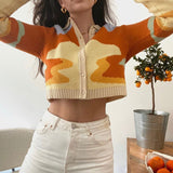 A woman wearing a cropped, collared cardigan with an abstract wave pattern in orange, yellow, and cream, paired with white jeans. A small potted tree with oranges is visible to the right.