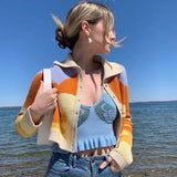 A woman wearing a cropped, collared cardigan with an abstract wave pattern in orange, yellow, and cream, over a blue ruffled crop top, paired with blue jeans. She is standing by the water.