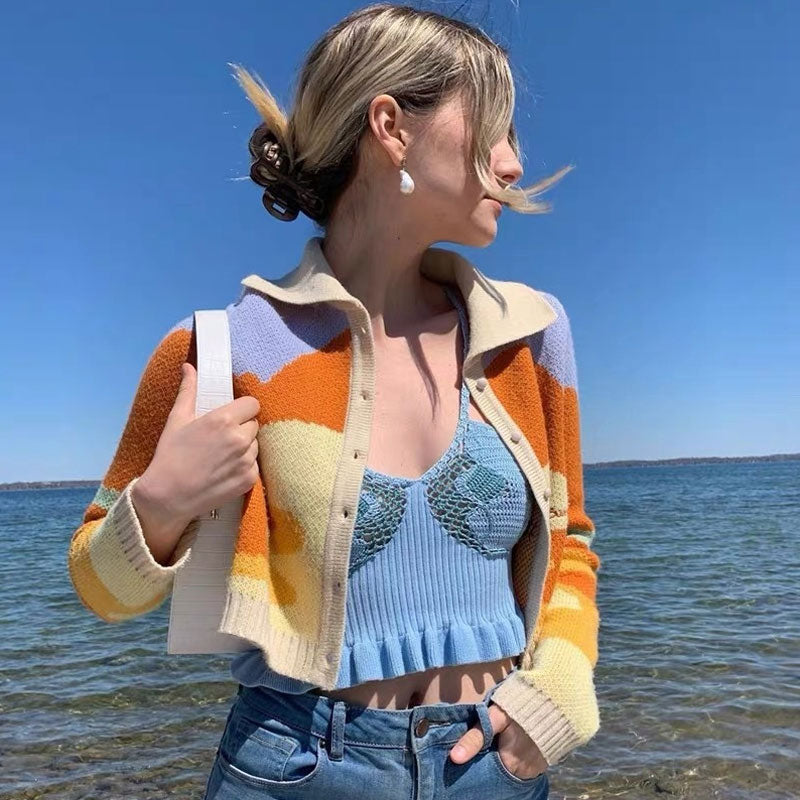 A woman wearing a cropped, collared cardigan with an abstract wave pattern in orange, yellow, and cream, over a blue ruffled crop top, paired with blue jeans. She is standing by the water.