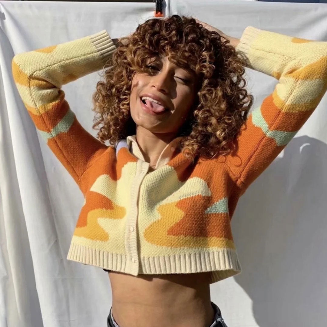 A woman with curly brown hair wearing a cropped, collared cardigan with an abstract wave pattern in orange, yellow, and cream. She has her hands behind her head and is sticking her tongue out.