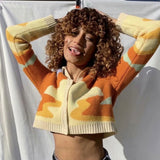 A woman with curly brown hair wearing a cropped, collared cardigan with an abstract wave pattern in orange, yellow, and cream. She has her hands behind her head and is sticking her tongue out.