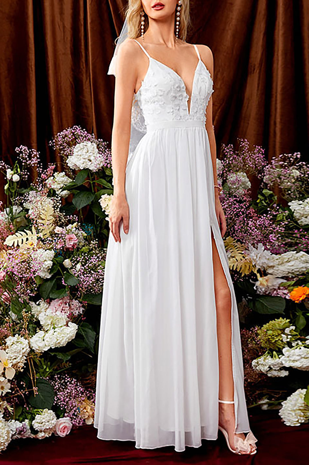 Woman in a white wedding dress with a deep V-neckline, floral appliques, and a high slit, standing in front of a floral backdrop.