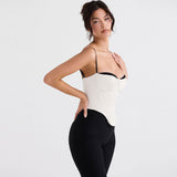 A woman wearing a white satin corset top with black trim and spaghetti straps, posing with hands on her hips.