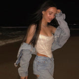A young woman with long dark hair wears a white strapless corset top, a denim jacket, and ripped jeans on a beach at night.