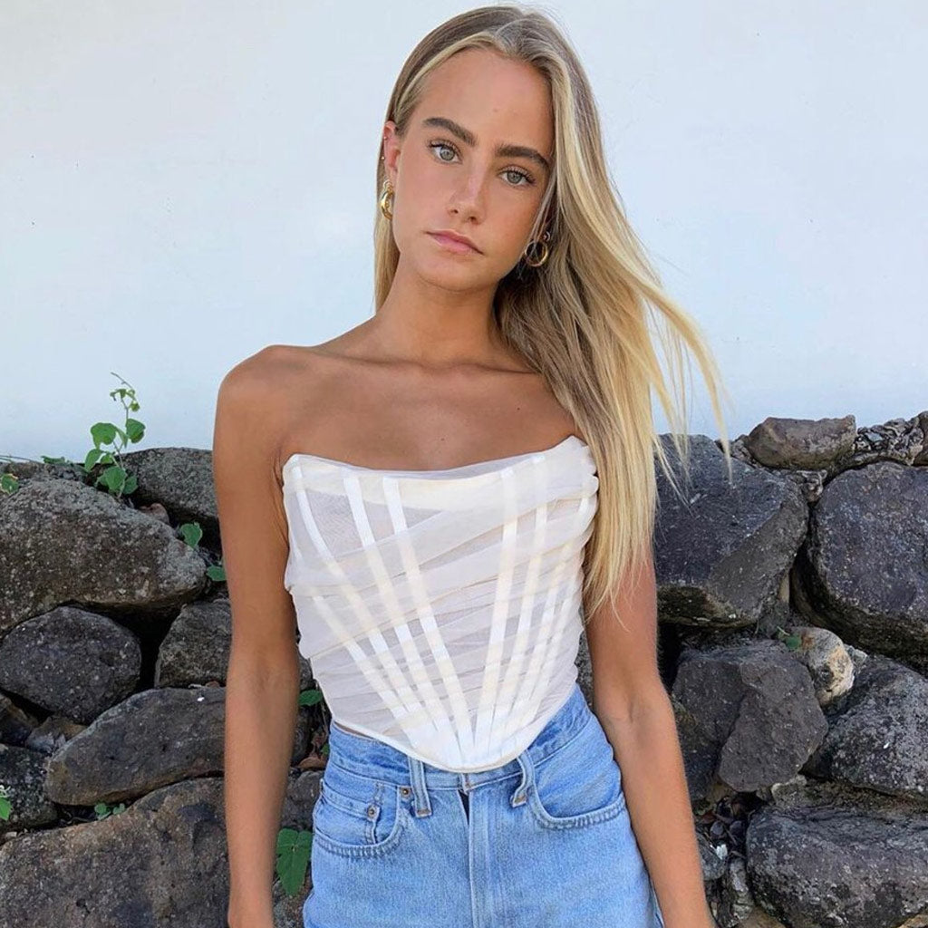 A young woman with blonde hair and blue eyes wears a white strapless corset top and blue jeans, posing against a stone wall.