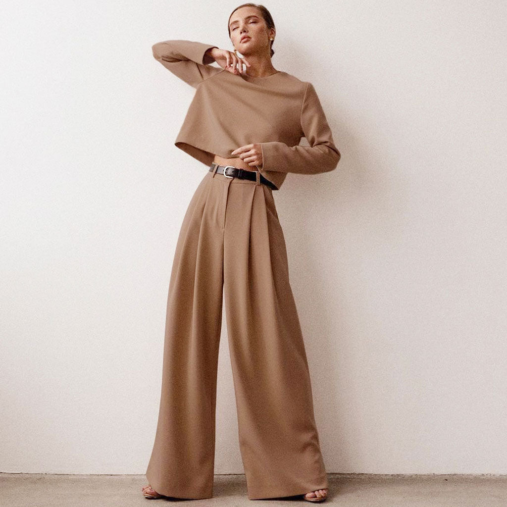 A woman in a tan wide-leg pant suit poses against a white wall.