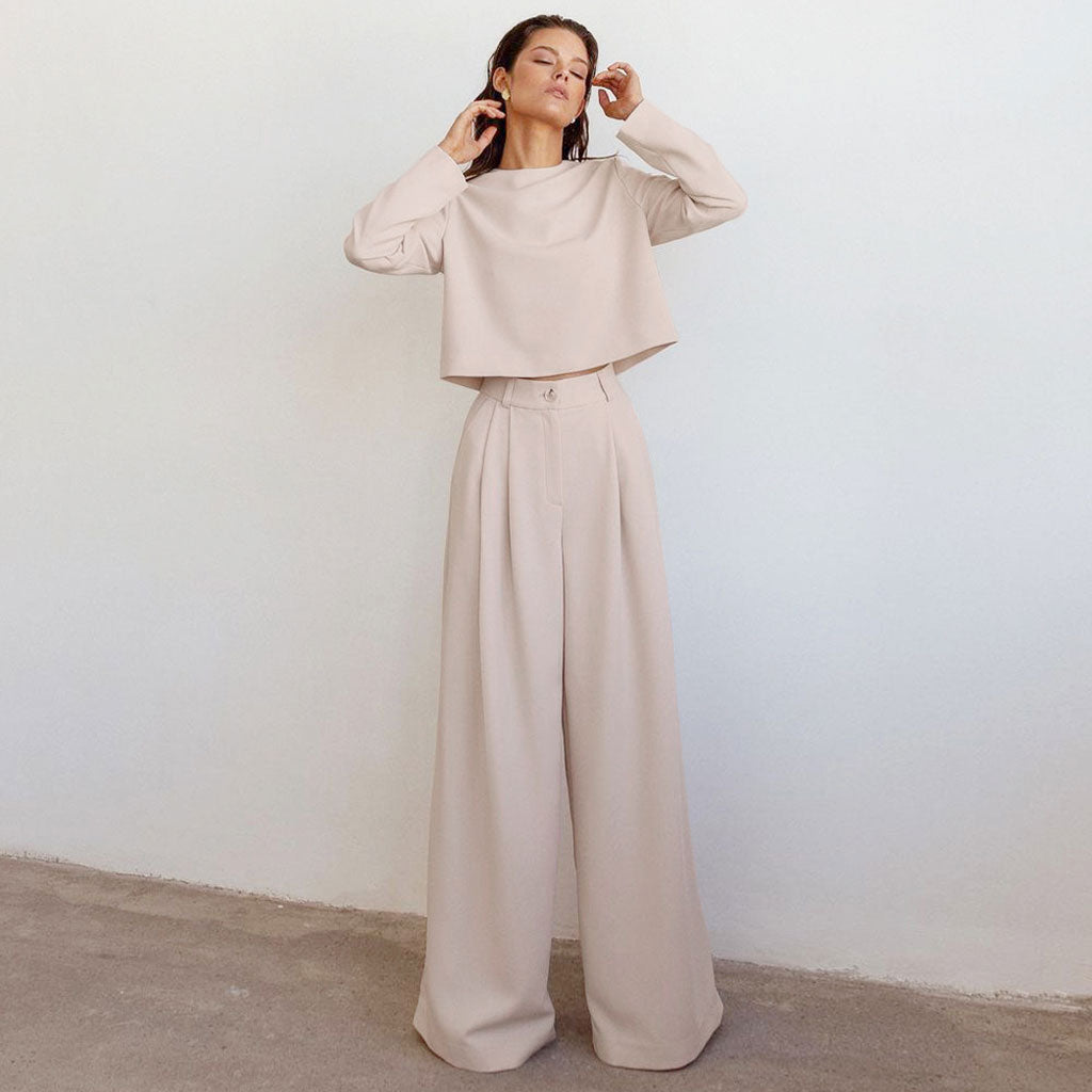 A woman in a beige crop top and wide-leg pants stands against a white wall.