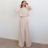 A woman in a beige crop top and wide-leg pants stands against a white wall.