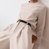 A close-up of a woman wearing a beige outfit with a brown belt, leaning against a wall.