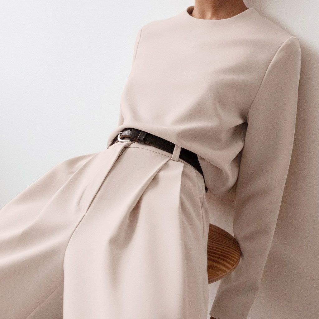 A close-up of a woman wearing a beige outfit with a brown belt, leaning against a wall.