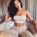 A woman with curly hair sits on a couch wearing a beige crop top and matching sweatpants.