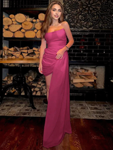 A woman in a fuchsia strapless dress with a draped asymmetrical hemline stands in front of a fireplace.