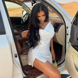 A woman in a white satin mini dress with a cowl neckline and a high slit looks down as she sits in the driver's seat of a white SUV in a desert setting.