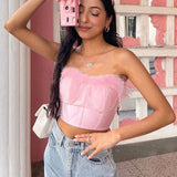 A young woman with long dark hair, wearing a pink strapless corset top with pink feather trim, denim jeans, and gold hoop earrings, holds a pink camera to her face.