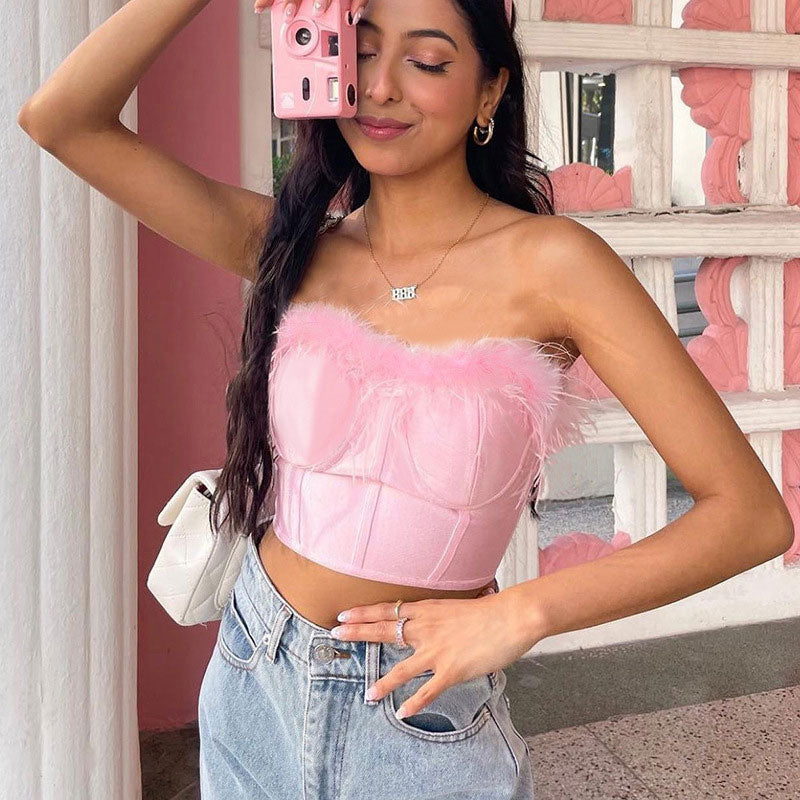 A young woman with long dark hair, wearing a pink strapless corset top with pink feather trim, denim jeans, and gold hoop earrings, holds a pink camera to her face.