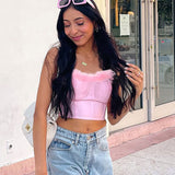 A young woman with long dark hair, wearing a pink strapless corset top with pink feather trim, denim jeans, and gold hoop earrings, smiles and touches her hair. She is wearing pink sunglasses.