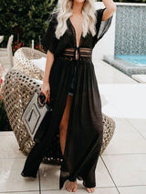Woman wearing a black sheer maxi dress with lace trim and a tie front, holding a patterned bag, standing by a pool.