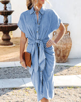 Woman wearing a light blue denim midi dress with short sleeves and a tie waist, holding a brown clutch.