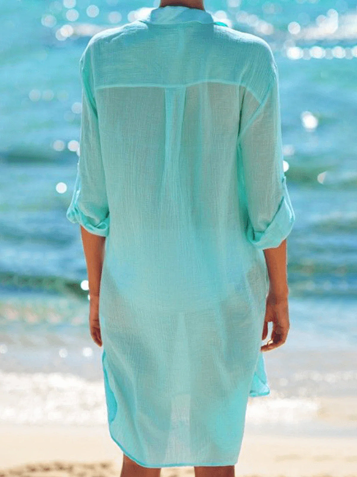 Back view of a woman wearing a mint green sheer button-down cover-up on a beach.