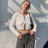 Woman in a white cropped cardigan and grey jeans leaning against a white brick wall.