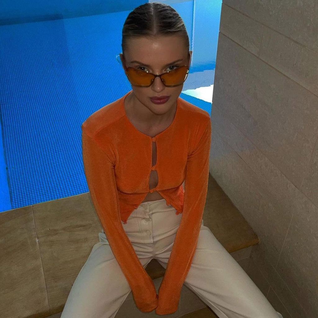 Woman wearing an orange cropped cardigan and white pants, sitting by a pool.