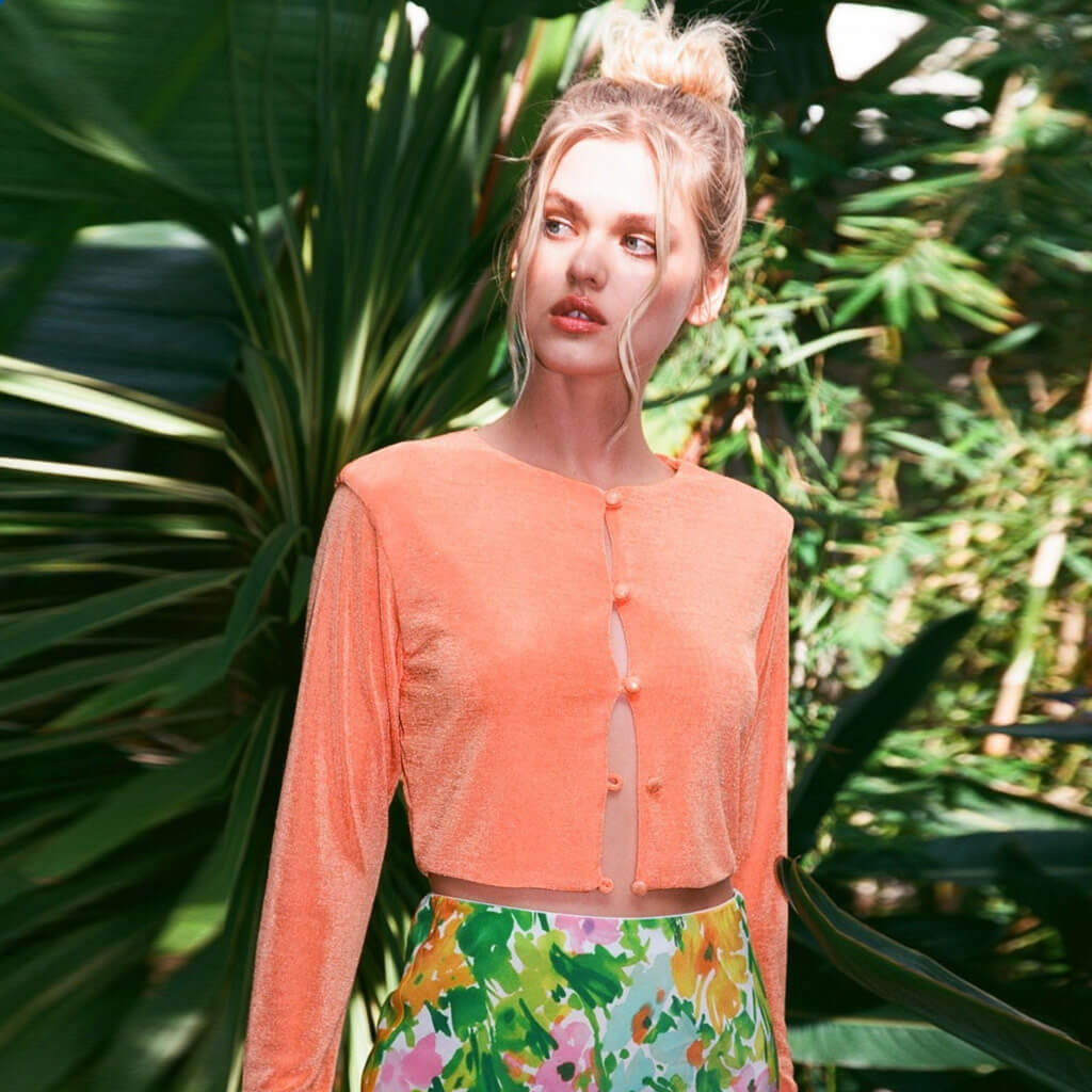 Woman with her hair in a bun wearing a peach cropped cardigan and floral pants in a tropical setting.