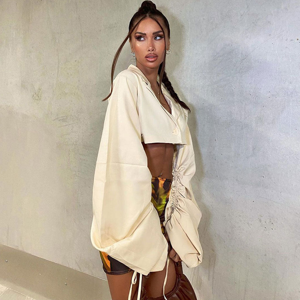 Woman wearing a cream cropped shirt with cinched sleeves and patterned shorts, posing against a concrete wall.