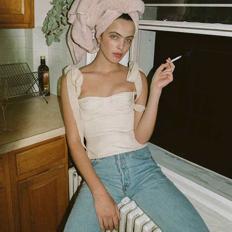 A young woman with a towel wrapped around her head sits on a windowsill, wearing a white ruched bustier top with tie shoulder straps and blue jeans, holding a cigarette.