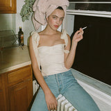 A young woman with a towel wrapped around her head sits on a windowsill, wearing a white ruched bustier top with tie shoulder straps and blue jeans, holding a cigarette.