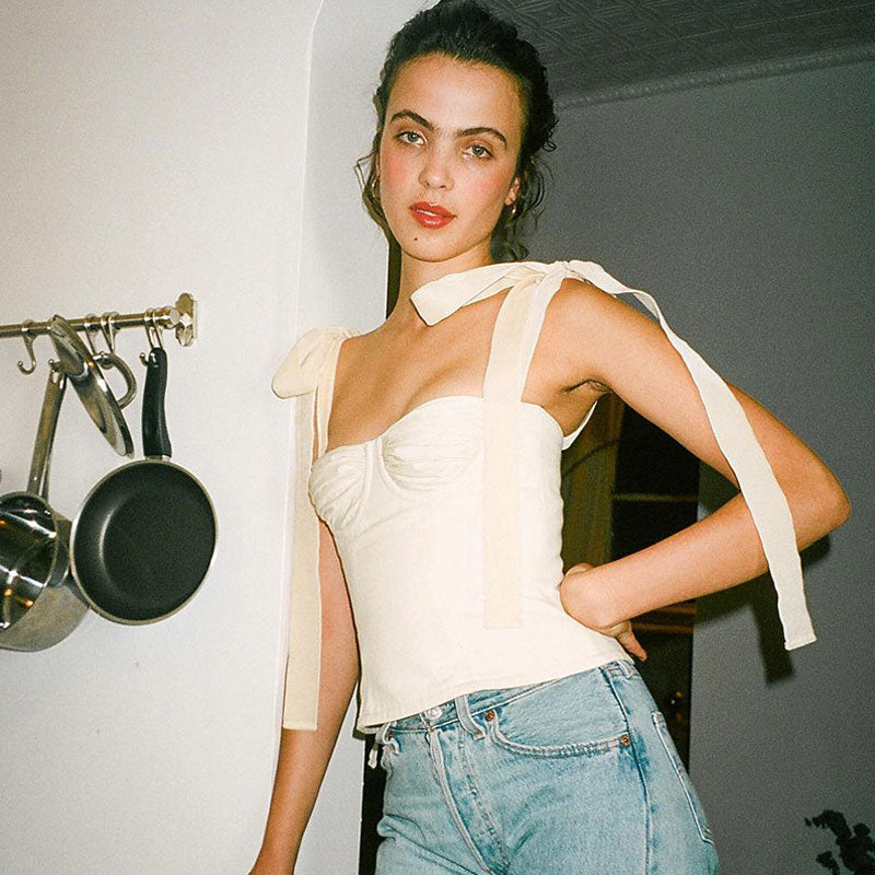 A young woman with her hair pulled back poses with her hand on her hip, wearing a white ruched bustier top with tie shoulder straps and blue jeans.