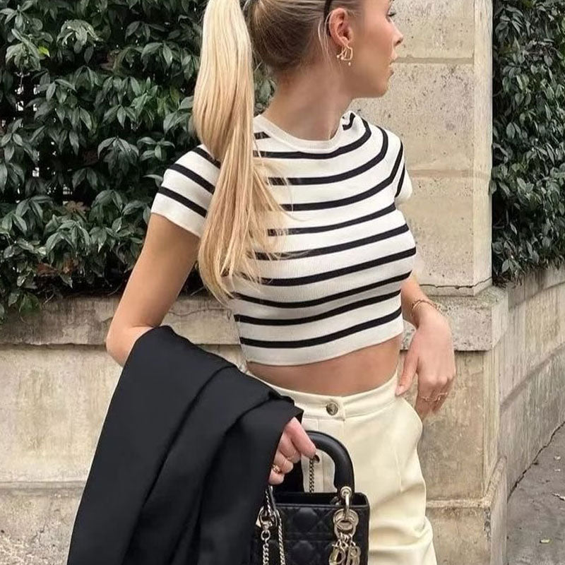 A woman with blonde hair in a ponytail wears a black and white striped crop top with a black blazer draped over her shoulder.