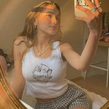 Young woman taking a selfie in a mirror, wearing a white ribbed tank top with lace trim and a graphic print of an angel and butterflies, paired with plaid shorts.