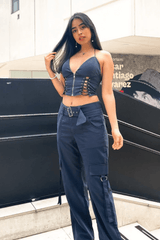 A young woman poses outdoors, touching her hair and looking at the camera in a navy blue lace-up crop top and cargo pants.