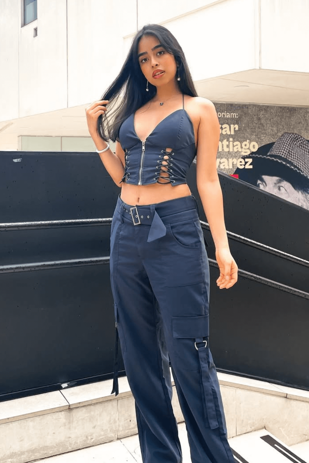 A young woman poses outdoors, touching her hair and looking at the camera in a navy blue lace-up crop top and cargo pants.