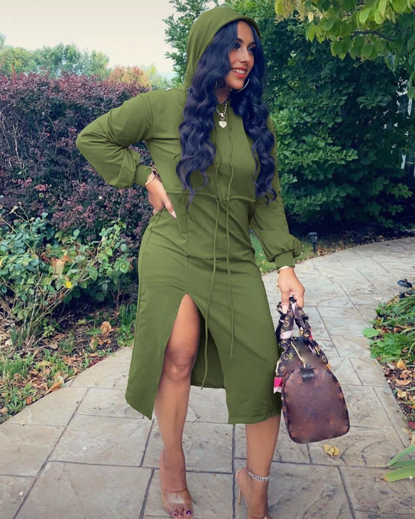 A woman in an olive green hooded midi dress with a high slit, clear heels, and hoop earrings, holding a designer handbag, standing outdoors on a paved path.