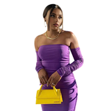 A woman wearing a purple off-the-shoulder ruched dress and holding a yellow purse.