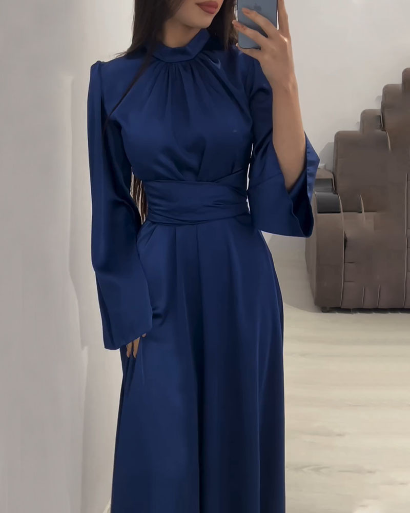 Woman wearing a long, navy blue satin dress with bell sleeves and a wide belt, standing in front of a mirror.