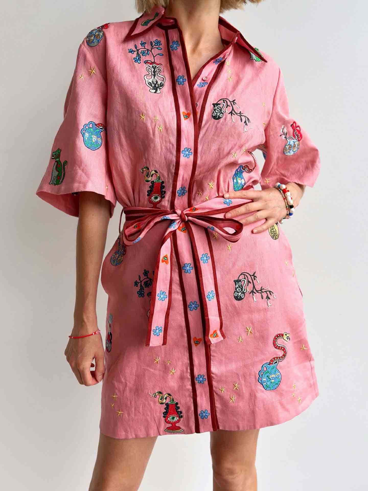 A woman in a pink shirt dress with colorful embroidered motifs and a matching belt, showing the front of the dress.