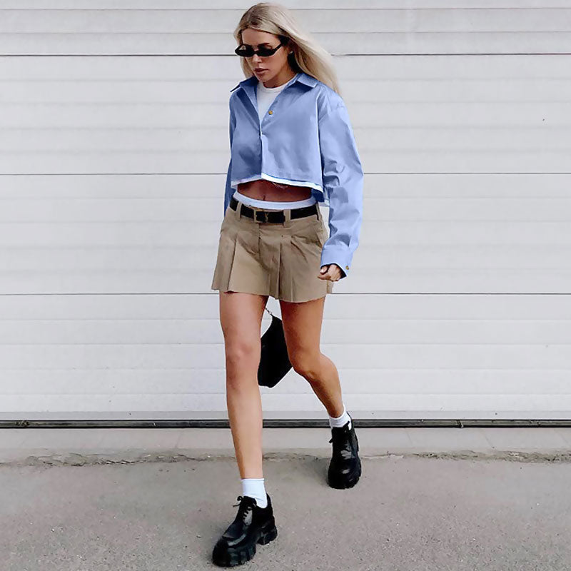 Full body shot of a woman in a light blue cropped button-down shirt, khaki pleated mini skirt, black chunky shoes, white socks, and black sunglasses, walking outdoors.