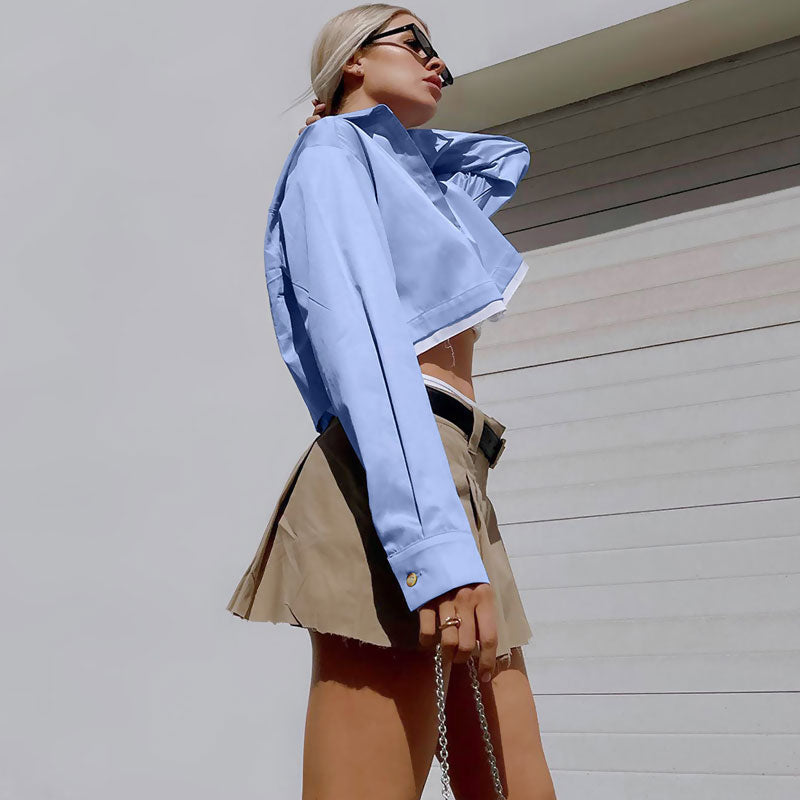 Close-up side profile of a woman wearing a light blue cropped button-down shirt and a khaki pleated mini skirt, with her hand touching her neck.