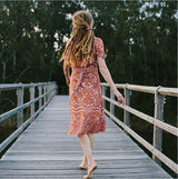 woman in orange floral wrap dress walking on a wooden bridge